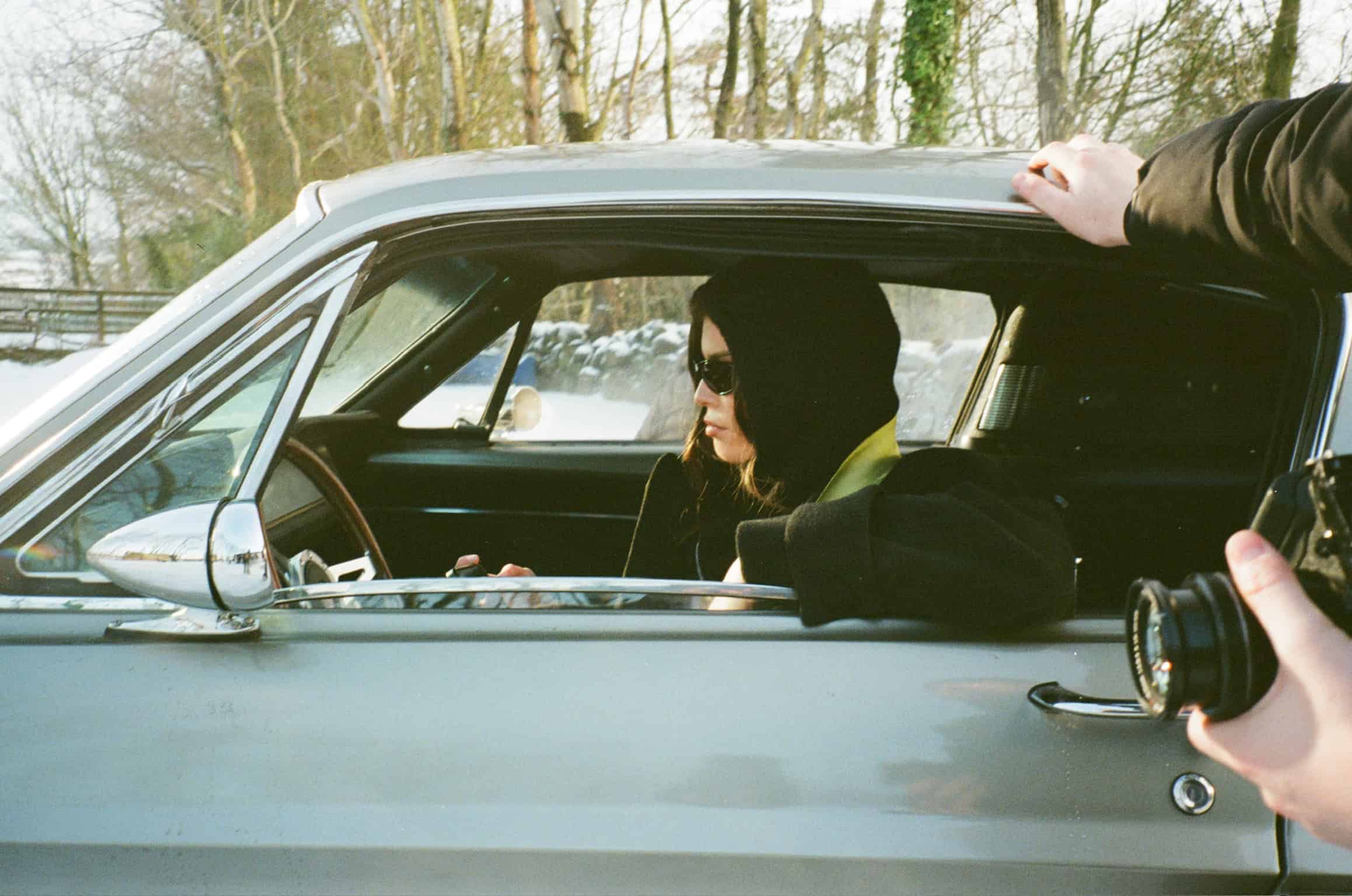 Campaign model Emma in the a vintage car, wearing the steal hoodie and the evergreen jacket, behind the scenes of the campaign photoshoot. Shot on 35mm colour film.