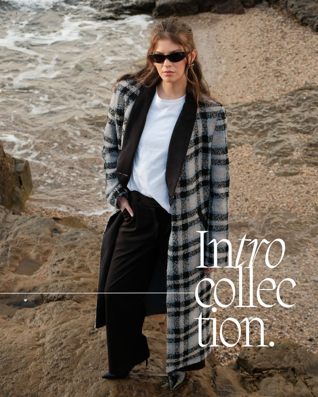 Female model Emma wearing the intro tshirt, harvest jacket and sonder suit trousers and sunglasses looking up at the camera from the shore of a coastal beach in Ireland.