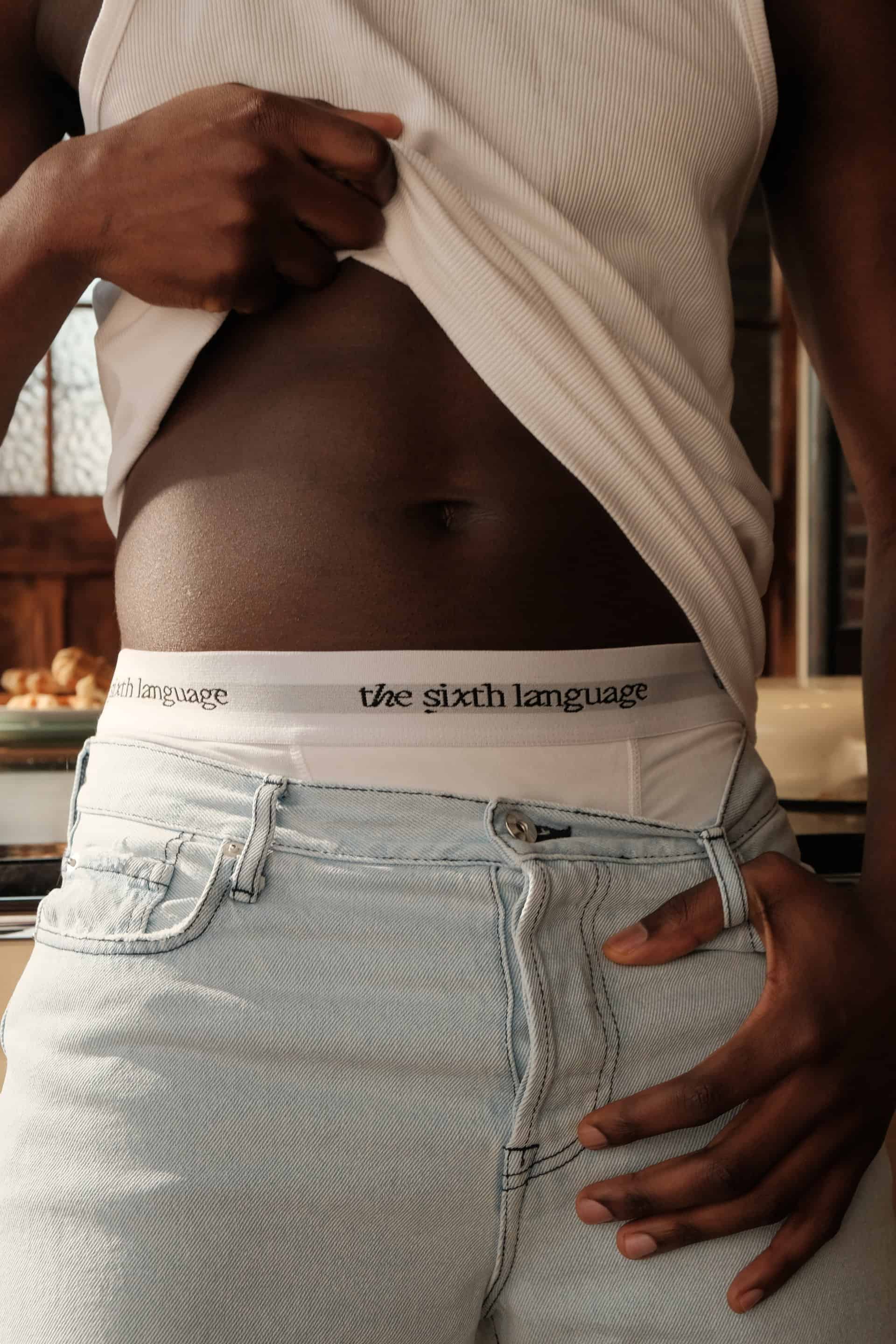 Male model patrick showing off "the roommates boxers" and "the backseat jeans"  in a close up shot, lifting his shirt up slightly to reveal the waistband showing the logo of The Sixth Language.
