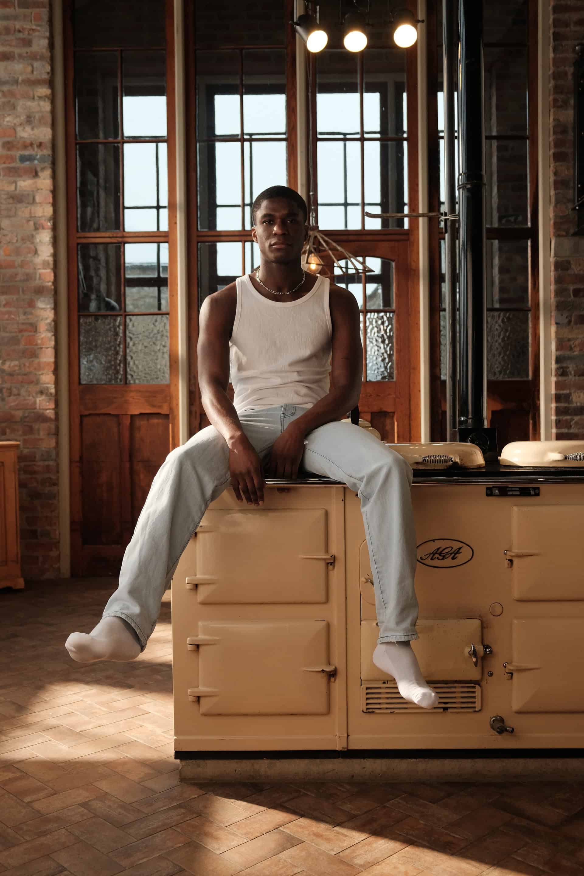 Male model Patrick sat casually on the stove and aga in a warm rustic kitchen, wearing the Sixth language's backseat jeans, wearing a vest and socks looking right at the camera