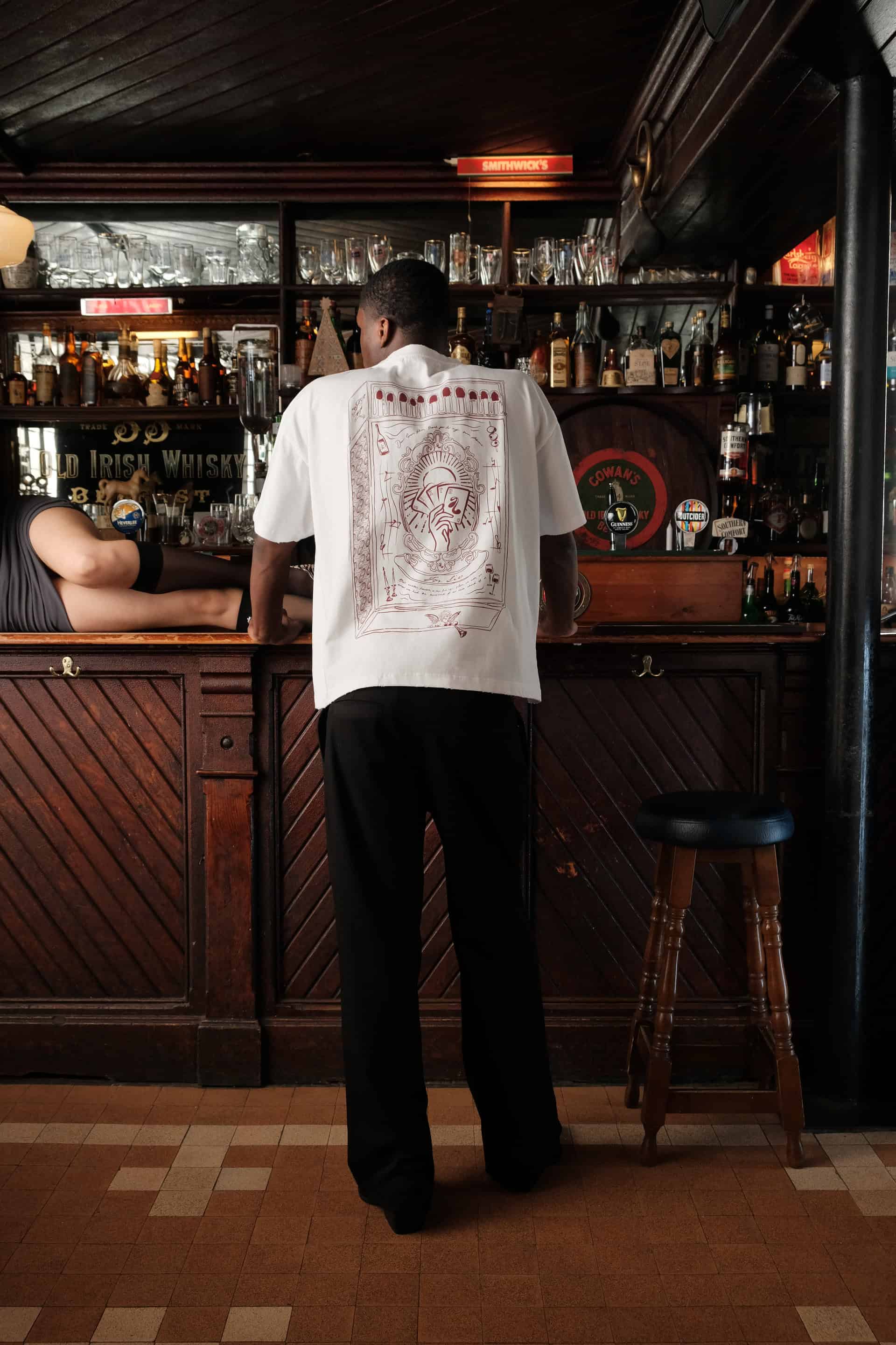 Male model Patrick standing at a bar wearing "the Intro Tshirt" and "Sonder Suit Trousers" faced away from the camera to see the intricate design on the rear of the tshirt, a statement piece of The Sixth Language's debut collection "the Intro Collection"