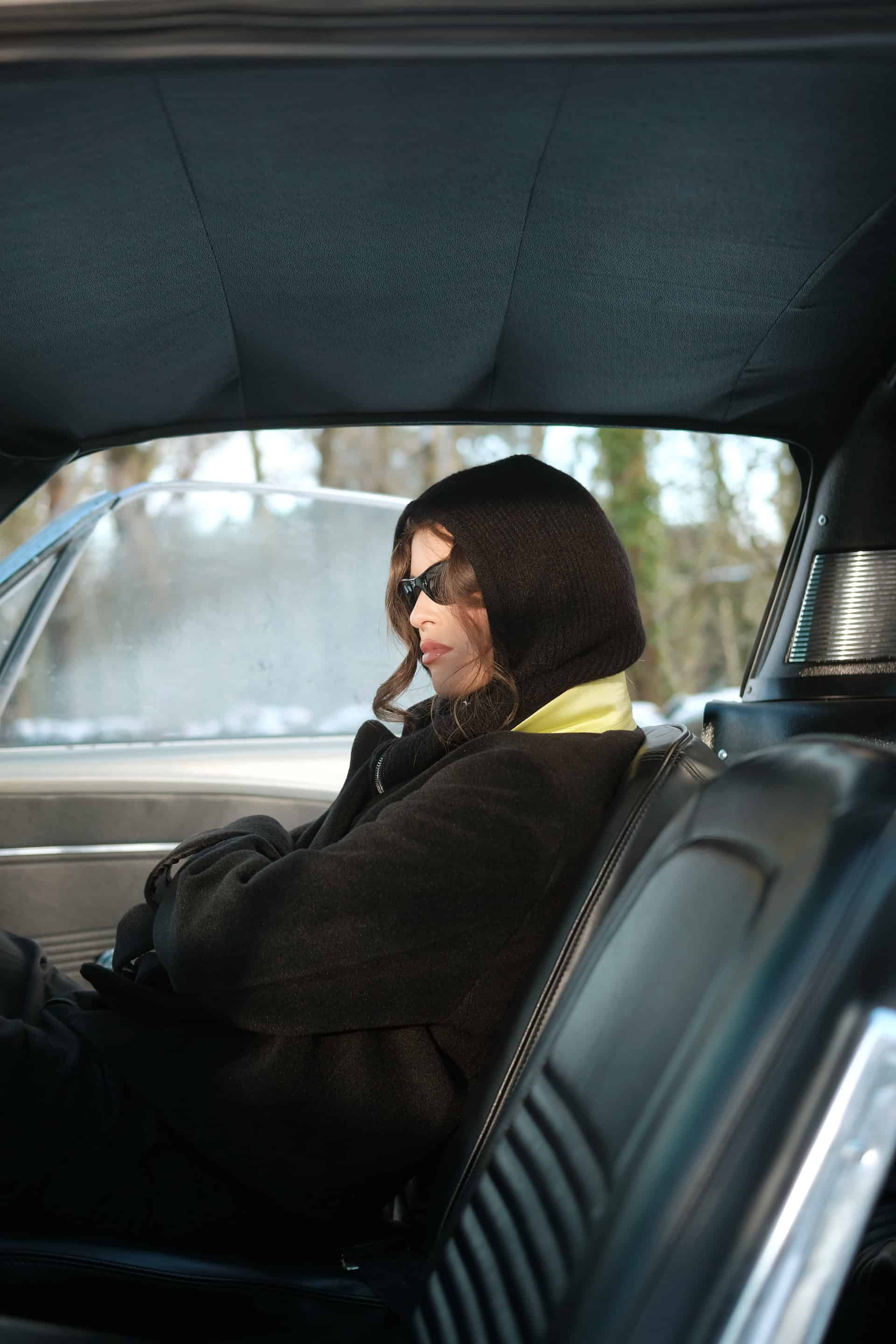 Female model Emma sitting in the vintage car looking distantly from the camera wearing the evergreen jacket and steal hoodie for The Intro Collection campaign.