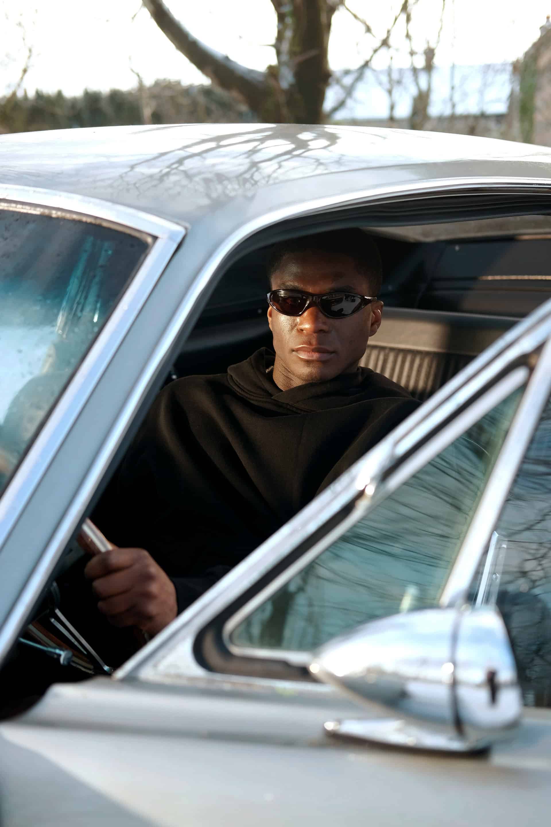 Model Patrick in the vintage car looking at the camera with sunglasses on through the body and door of the car, wearing The Steal Hoodie of the Sixth Languages debut collection.