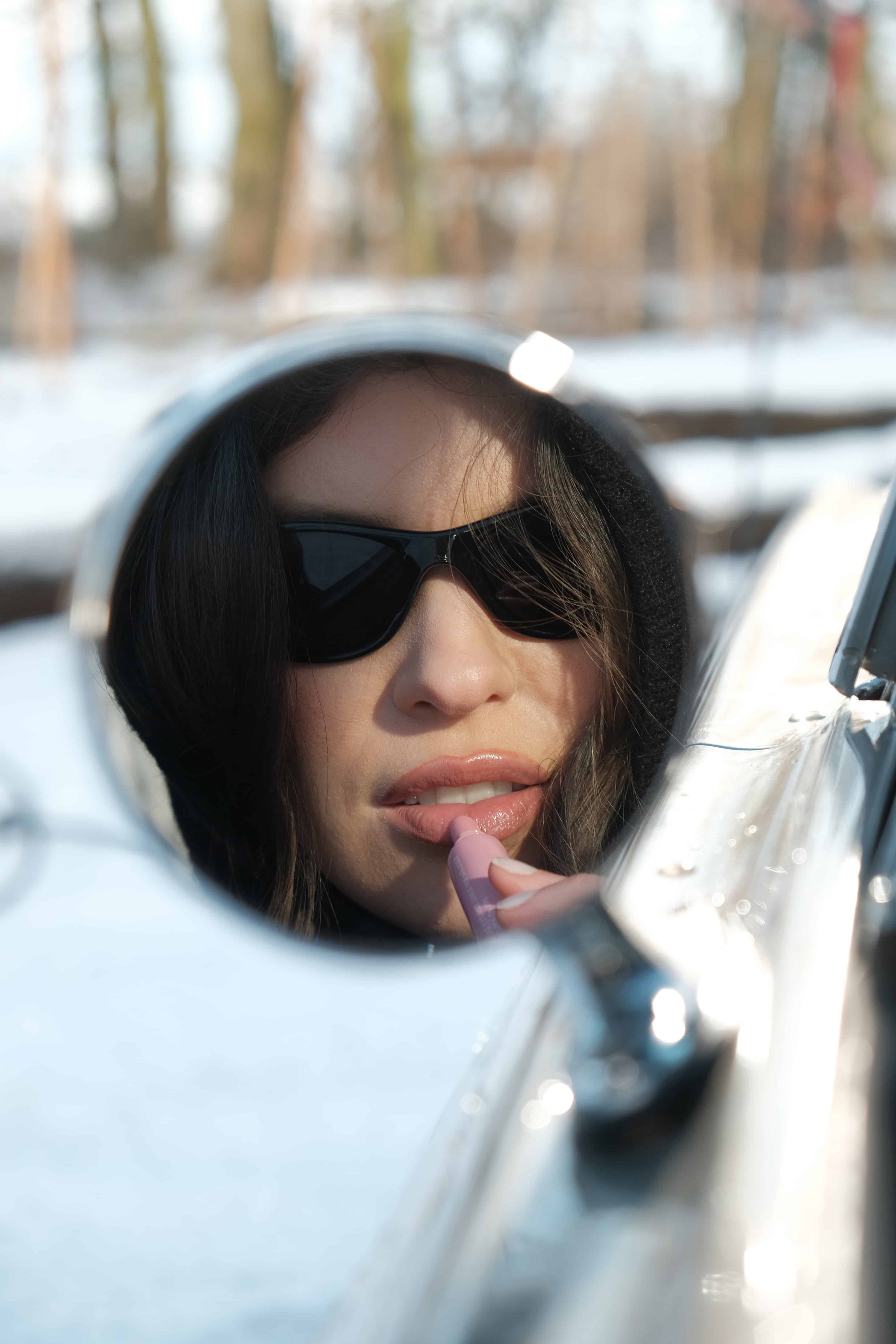 The reflection of Emma looking in the wing mirror of a vintage car putting on lipstick.