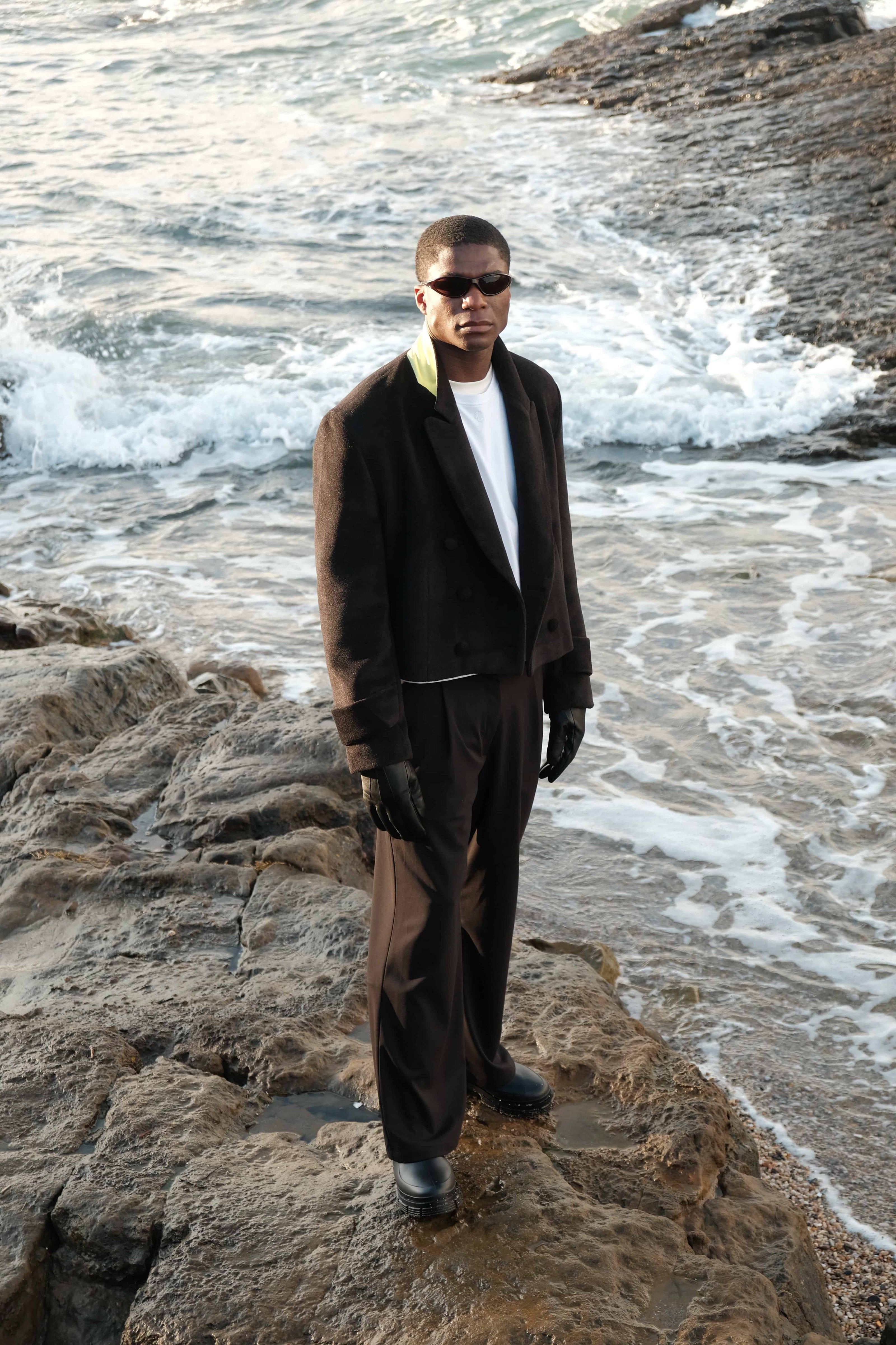 Male model Patrick standing on the rocks on the shore of a coastal beach of ireland, wearing the evergreen jacket, sonder suit trousers and masterpiece t-shirt - looking up into the camera.