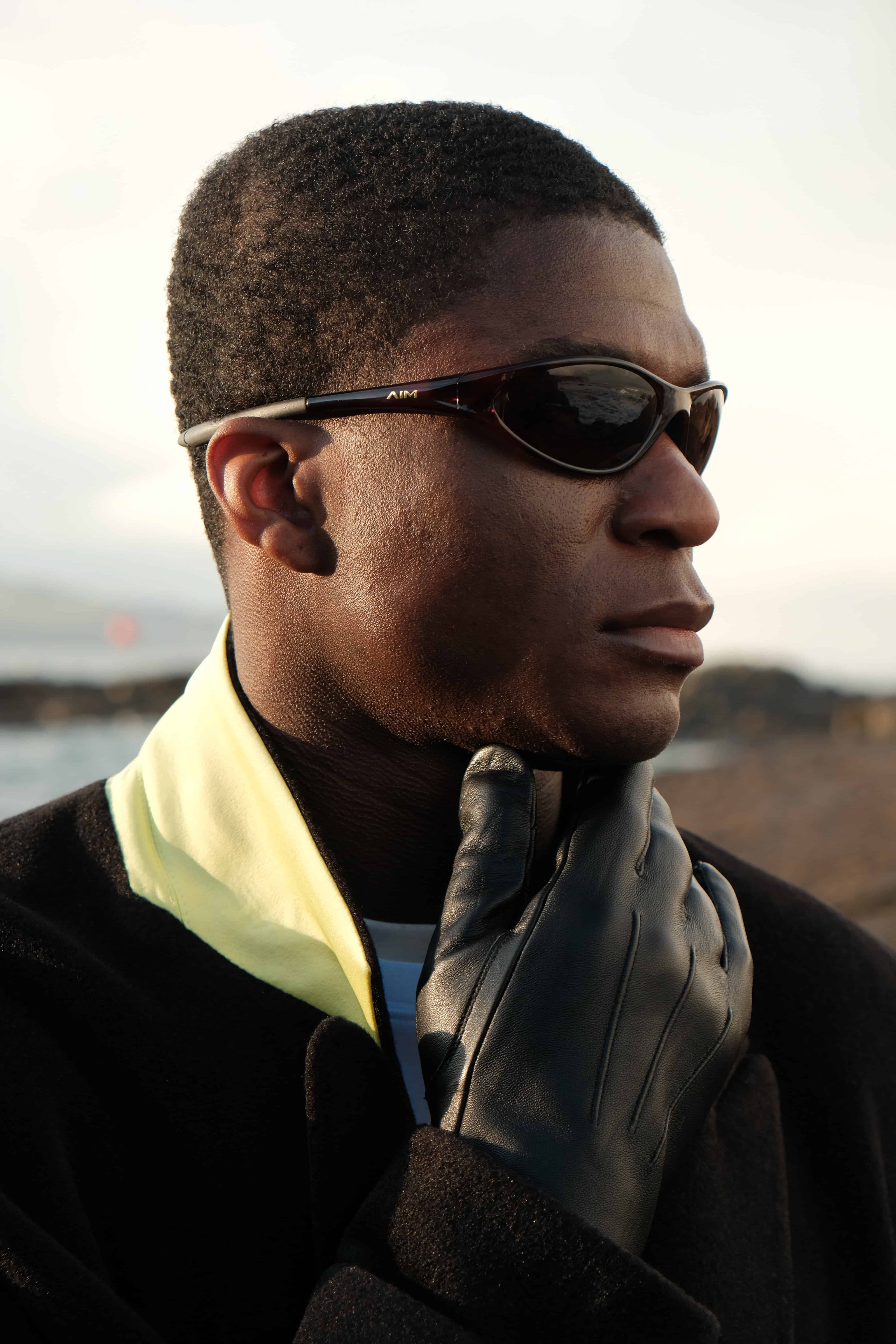 Close-up profile of Patrick looking away from the camera, wearing the evergreen jacket, leather glove and sunglasses looking into the middle distance.