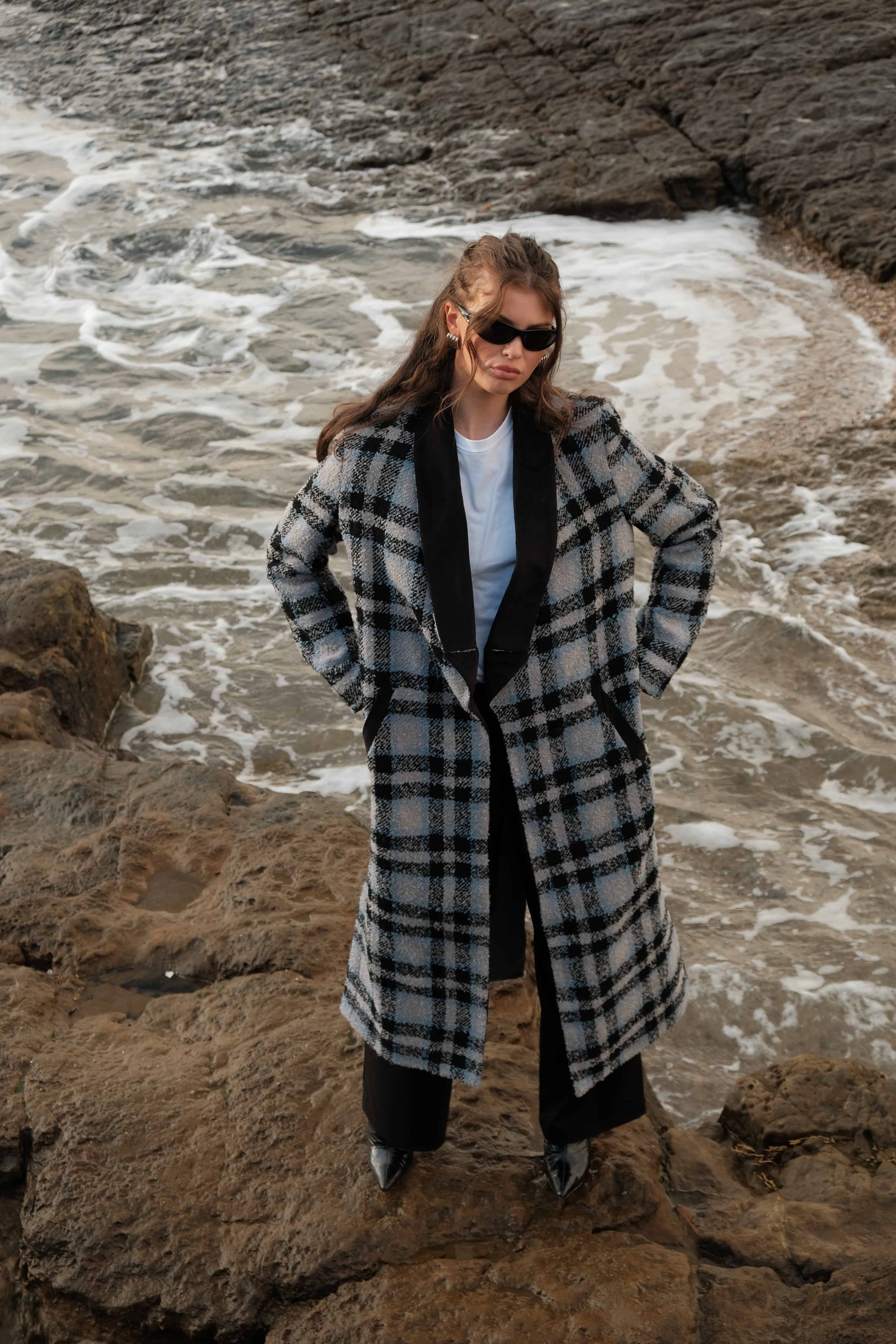 Female model Emma wearing the intro tshirt, harvest jacket and sonder suit trousers and sunglasses looking up at the camera from the shore of a coastal beach in Ireland, with her hands on her sides.